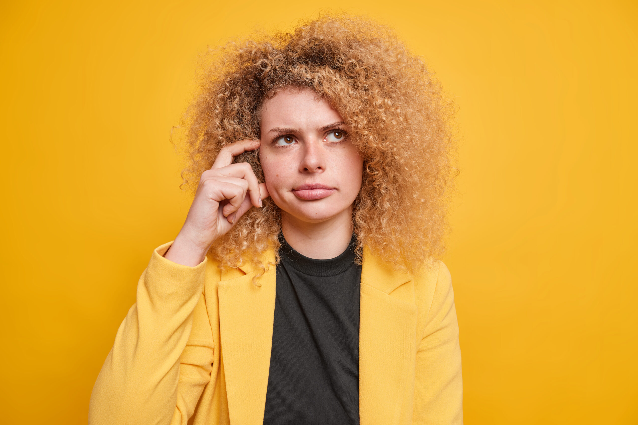Portrait of pensive displeased curly female entrepreneur in formal clothes scratches temples tries to remember important information comes up with ideas poses indoor against yellow background