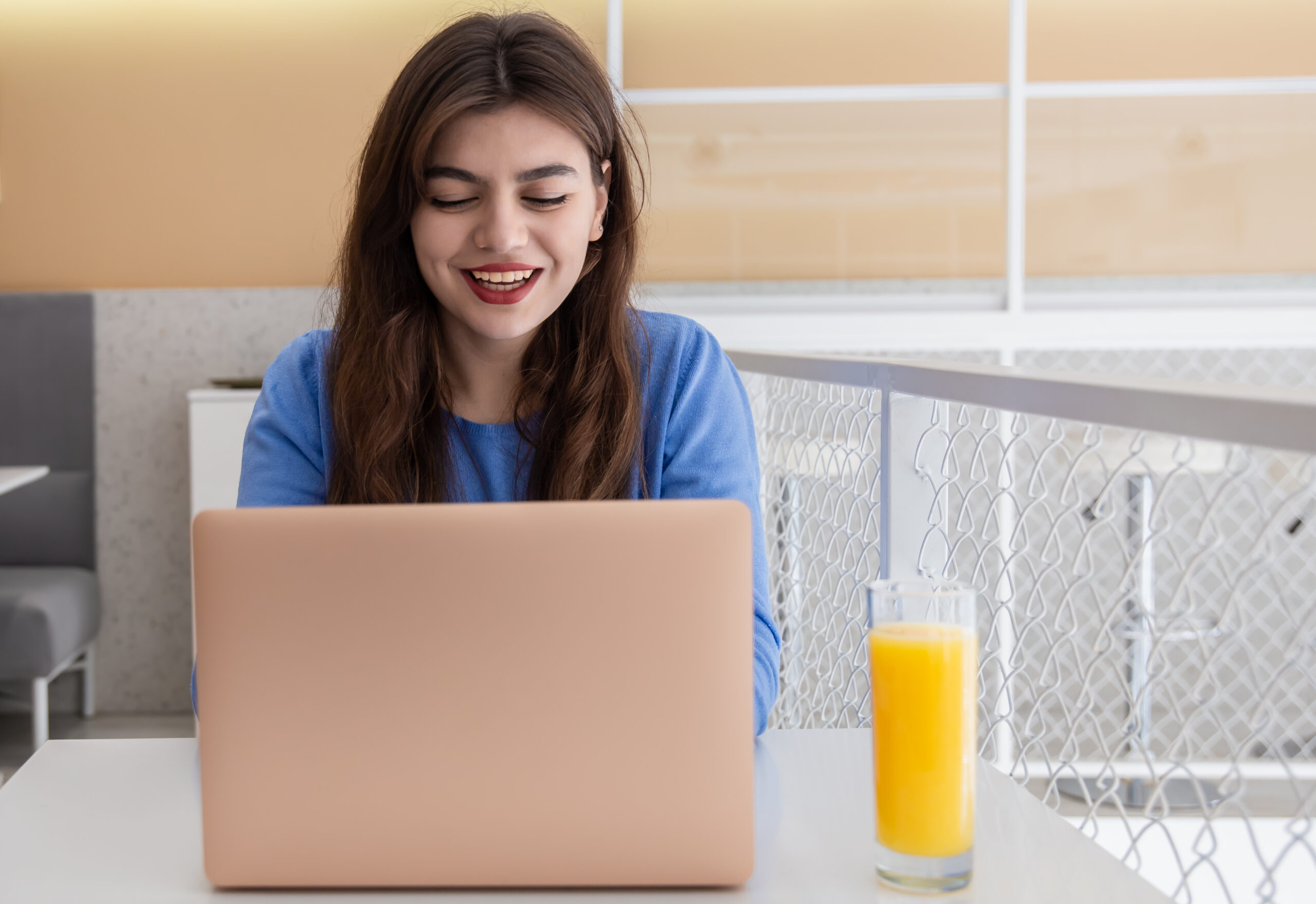 A young woman with an attractive smile, red lipstick, in a blue sweater works at a laptop, sitting in a cafe.