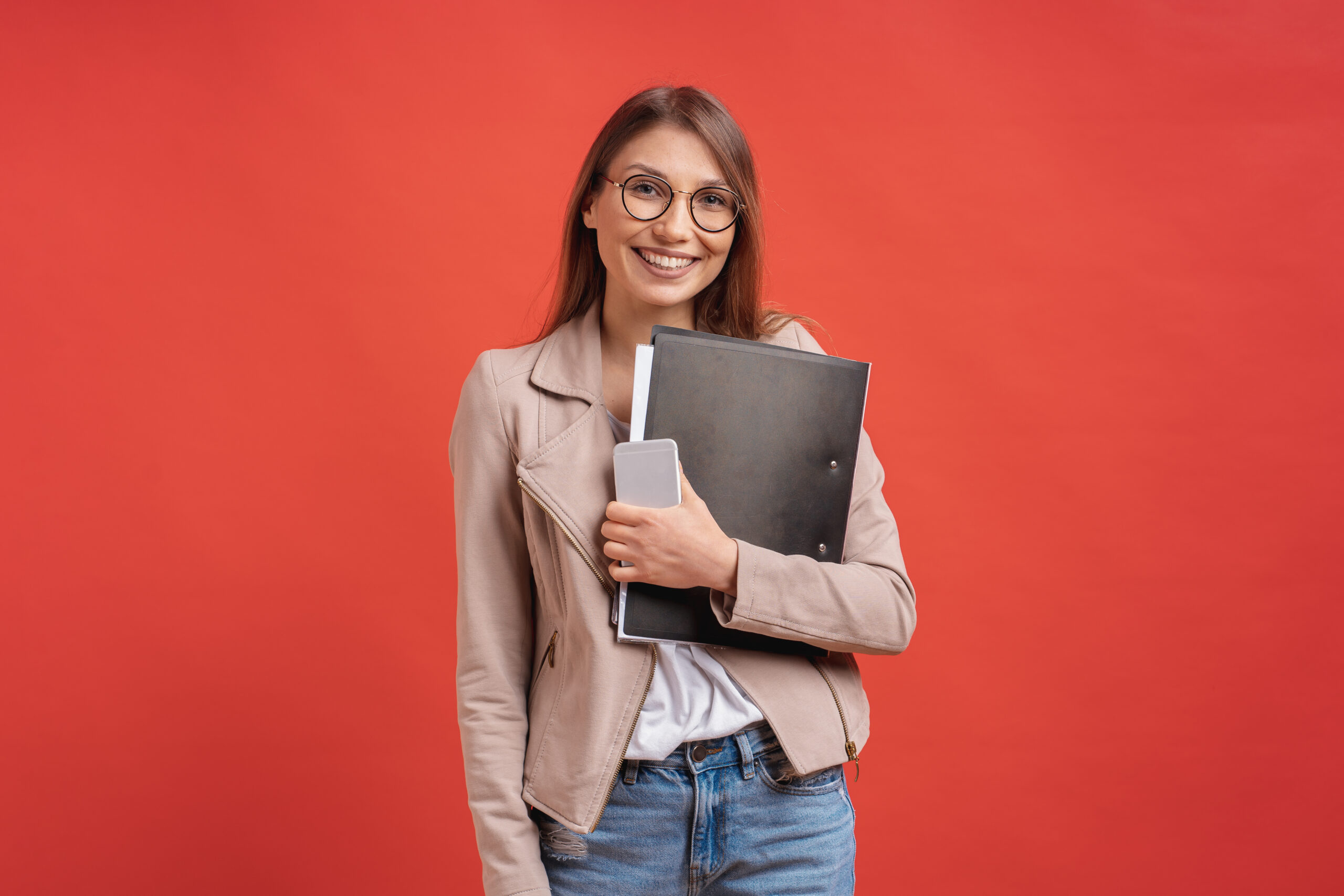 Young smiling student or intern in eyeglasses standing with a folder in hands and happily looking to camera over red background in the studio.