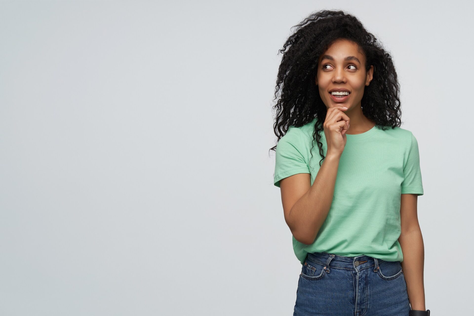 Happy pensive african american young woman with long curly hair in mint tshirt keeps hand on chin and thinking isolated over gray background